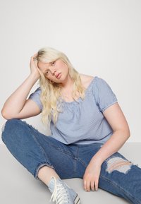 Blusa azul e branca às riscas, com decote off-the-shoulder e gola franzida, combinada com jeans azuis desgastados e ténis azul claro.