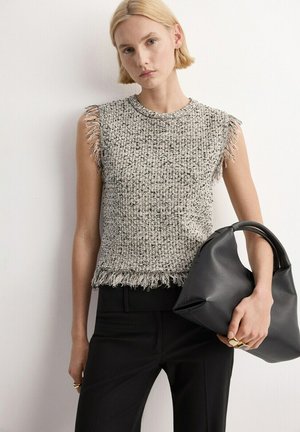 Mujer rubia con un top gris de tweed sin mangas con flecos y pantalones negros, sosteniendo un bolso de cuero negro de estilo relajado a su lado.