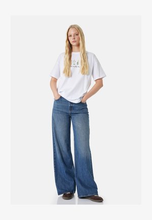 Jeune femme aux longs cheveux blonds portant un t-shirt blanc à motif floral, un jean bleu à jambes larges et des chaussures marron, debout les mains dans les poches.