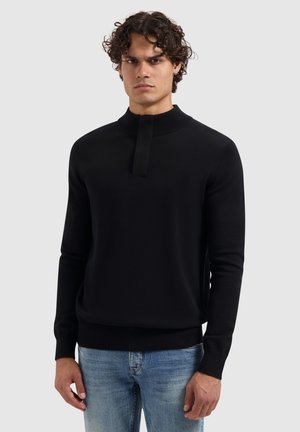 Mann mit lockigem Haar, der einen schwarzen Pullover mit langen Ärmeln und hohem Kragen sowie hellblauen Jeans trägt, steht vor einem schlichten Hintergrund.