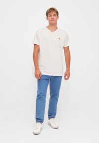 Kortærmet beige V-hals t-shirt med rødt logo, parret med lyseblå denimjeans og hvide sneakers. Simpel tekstur, afslappet design.