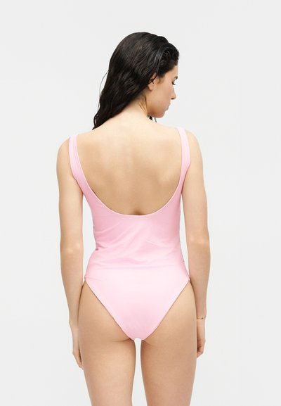 Femme aux longs cheveux bruns portant un maillot de bain une pièce rose avec un dos échancré, debout de dos devant un fond uni.