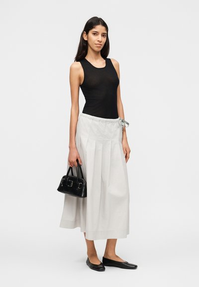 Γυναίκα που φοράει μαύρο αμάνικο τοπ, λευκή φαρδιά φούστα με κορδόνι, μαύρες μπαλαρίνες, κρατώντας μια μικρή μαύρη τσάντα, στέκεται σε λευκό φόντο.