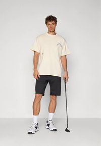 Beež lühikeste varrukatega T-särk tekstiga "STUDENT", mida kannavad mustad sportlikud lühikesed püksid, valged sokid ja valged tossud, hoides kämblasse golfikeppi.