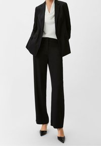 Blazer nero su misura e pantaloni a gamba larga con una maglia bianca a cascata. Texture liscia, design strutturato e décolleté neri a punta.