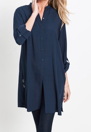 Kobieta w długiej granatowej koszuli na guziki z podwiniętymi rękawami i czarnych legginsach, pozująca z jedną ręką przy twarzy.