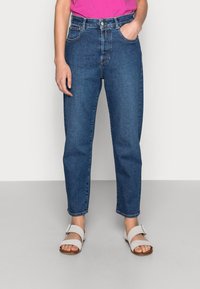 Jeans de mezclilla azul de talle alto con pierna recta, que cuentan con bolsillos frontales y cierre de botones. Combinados con sandalias blancas de tiras.