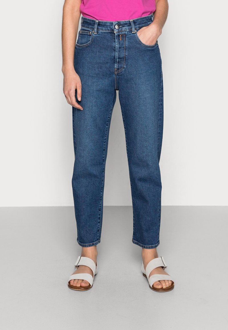 Jeans de mezclilla azul de talle alto con pierna recta, que cuentan con bolsillos frontales y cierre de botones. Combinados con sandalias blancas de tiras.