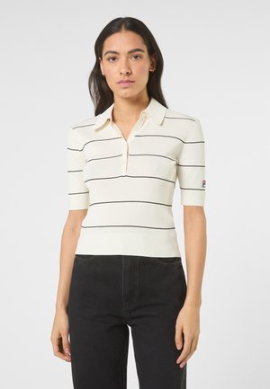 Giovane donna che indossa una polo color crema a maniche corte con sottili righe orizzontali nere e pantaloni neri su sfondo neutro.