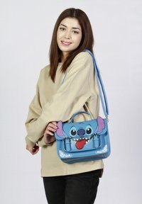 Bolsa sintética azul con un personaje de dibujos animados que tiene grandes orejas y una expresión juguetona. El diseño incluye acentos florales y una correa desmontable.