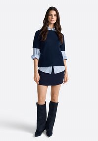 Un pull surdimensionné bleu marine superposé sur une chemise rayée bleu clair, associé à une jupe ajustée bleu marine et des bottes montantes à rayures.