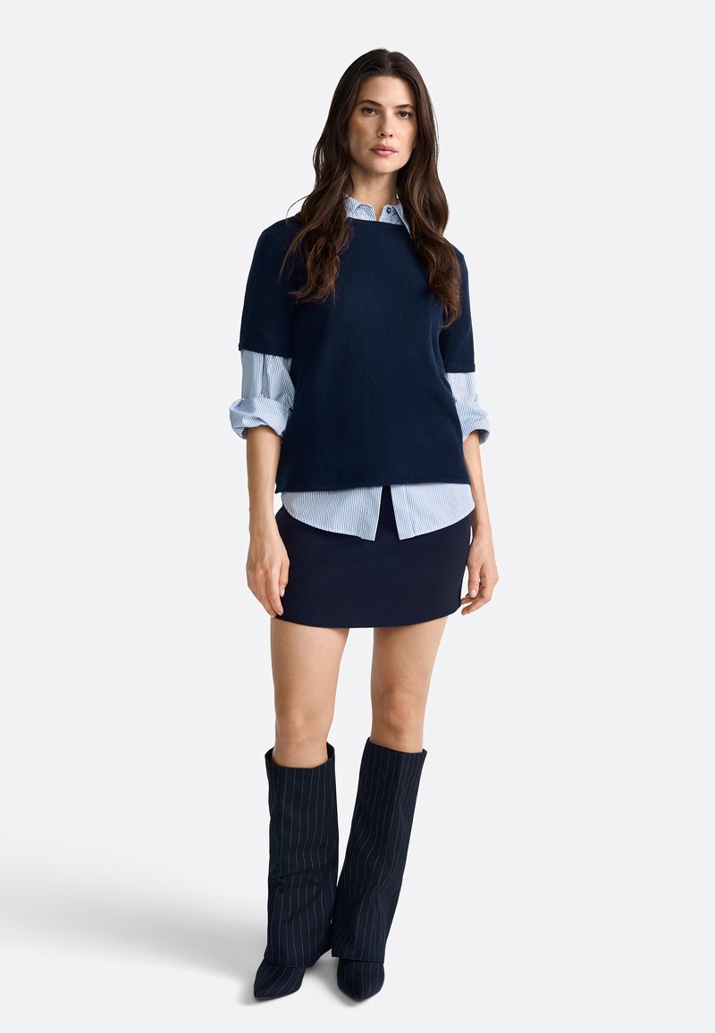 Un pull surdimensionné bleu marine superposé sur une chemise rayée bleu clair, associé à une jupe ajustée bleu marine et des bottes montantes à rayures.