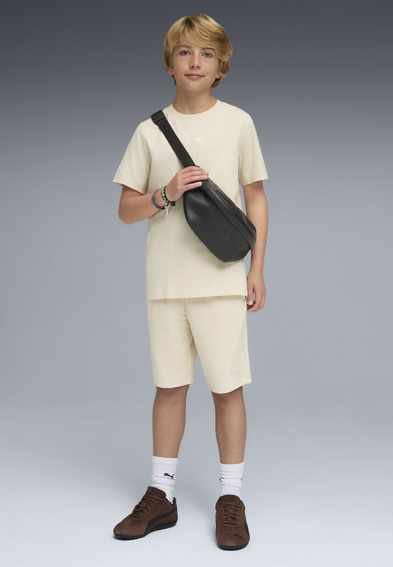 Garçon blond portant un t-shirt crème Puma et un short, tenant un sac bandoulière noir, avec des chaussures Puma marron et des chaussettes blanches sur un fond gris.
