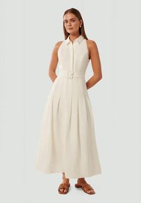 Vestido midi sem mangas, com cinto, em creme claro, com parte frontal de botões, saia plissada e detalhe de colarinho. Usado com sandálias castanhas.