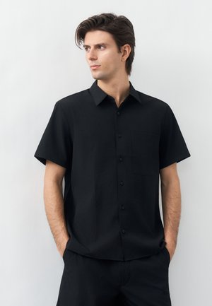 Młody mężczyzna o ciemnych włosach, ubrany w czarną krótką koszulę na guziki i czarne spodnie, stoi z rękami w kieszeniach, patrząc w lewo.