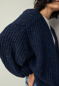 Cardigan en maille bleu marine avec une coupe ample et oversize, doté d'un tissu texturé et d'un devant ouvert. Le modèle porte un jean en denim.