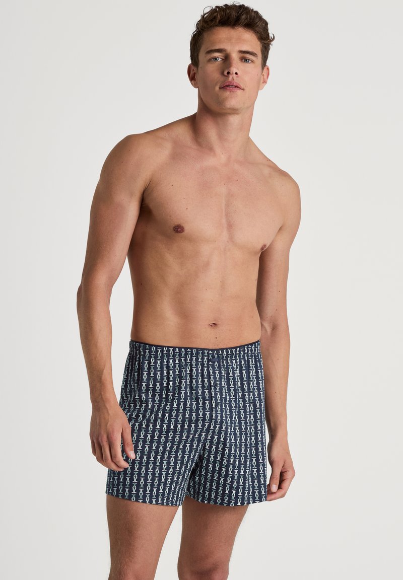 Herenboxers in donkerblauw met een herhalend wit patroon van abstracte vormen. De stof lijkt lichtgewicht en heeft een elastische tailleband.