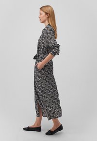 Schwarzes Maxikleid mit floralen Mustern, langen Ärmeln, geraffter Taille, Seitenschlitzen und strukturiertem Stoff. Flache schwarze Schuhe runden das Outfit ab.