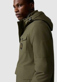 Giacca esterna verde oliva con cappuccio, molteplici tasche e una patch sul petto. Presenta una texture liscia e un design strutturato.