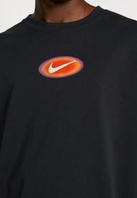 Musta puuvillapaita, jossa on pyöreä, monivärinen logo, jossa on Nike-viiva valkoisena, keskittyneenä rintaan. Sujuva pinta.