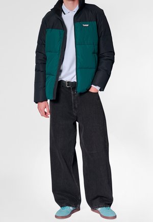 Mand iført sort og grøn dunjakke, hvid polo, brede sorte jeans, sort bælte og turkise sneakers mod en hvid baggrund.