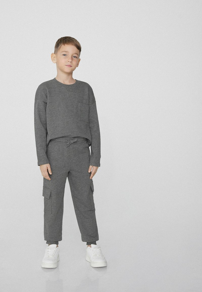 Szara bluzka z długim rękawem z kieszenią, zestawiona z pasującymi spodniami typu cargo z bocznymi kieszeniami i elastycznym sznurkiem w talii, uzupełniona białymi trampkami.