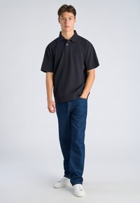 Jonge man die staat, gekleed in een zwart poloshirt met korte mouwen, een blauwe spijkerbroek en witte sneakers, met één hand in zijn zak tegen een effen lichte achtergrond.