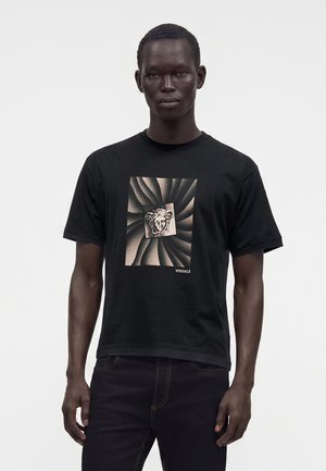 Bărbat purtând un tricou negru Versace cu un model învârtit și logo cu capul Meduzei în centru, asortat cu pantaloni negri.