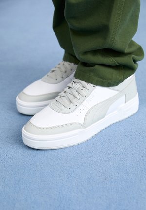 Persona con zapatillas Puma blancas y gris claro con cordones grises y pantalones verdes, de pie sobre un suelo con alfombra azul.