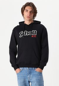 Felpa nera con cappuccio con logo "Schott NYC" in bianco e rosso. Presenta una tasca a marsupio e polsini a coste. Indossata con jeans azzurri.