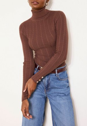 Frau trägt einen enganliegenden braunen gerippten Rollkragenpullover mit goldenen Knöpfen an den Manschetten und hoch taillierte blaue Jeans mit einem braunen Gürtel.