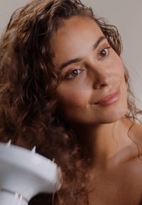 Witte föhn met een puntige mondstuk, gehouden in de buurt van golvend, bruin haar. Model met een gemiddelde huidskleur toont een natuurlijke make-uplook en zachte gelaatstrekken.