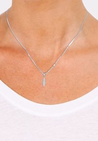 Collier pendentif en forme de plume argentée porté autour du cou d'une personne sur une chemise blanche, gros plan montrant la peau et la zone de la clavicule.