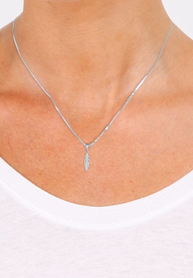 Collier pendentif en forme de plume argentée porté autour du cou d'une personne sur une chemise blanche, gros plan montrant la peau et la zone de la clavicule.