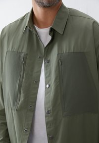 Olive green shirt jacket made of lightweight fabric, featuring two large zippered pockets, snap buttons, and a plain white T-shirt underneath.