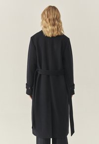 Tume mere villane mantle, millel on vööga talje, pikad varrukad ja minimalistlik disain. Omab selja lõhikut ja nööpidega mansette.