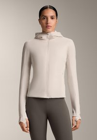 Ženska v oprijeti bež jakni z zadrgo, kapuco in odprtinami za palce, v kombinaciji s temnimi hlačami z visokim pasom, stoji pred nevtralnim ozadjem.