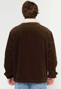 Mand iført en brun kordfløjljakke med krave af hvid fleece, set fra bagfra, parret med lyseblå jeans.