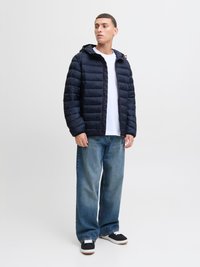 Giacca puffer blu navy con design trapuntato, chiusura con zip e cappuccio, abbinata a jeans a gamba larga blu chiaro e sneakers nere con accenti bianchi.