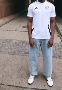 Jersey blanca de Adidas con acentos negros, escudo de Alemania; combinada con jeans de pierna ancha azul claros y zapatillas blancas con rayas negras.