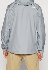Harmaa vedenpitävä takki hupulla, joustavilla hihojen resoreilla ja logolla yläselässä. Virtaviivainen muotoilu, sileä pinta ja kevyt materiaali.