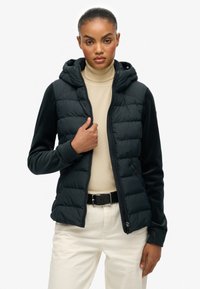 Veste matelassée noire avec capuche, présentant un devant en tissu lisse et des manches douces. Fermeture éclair à l'avant et poches latérales visibles.