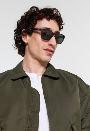 Giovane uomo con capelli ricci che indossa occhiali da sole neri, una camicia bianca e una giacca verde oliva con cerniera, che guarda di lato.