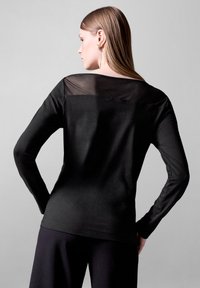 Maglietta nera a maniche lunghe con un pannello trasparente sulla parte superiore della schiena. Realizzata in un tessuto morbido con un design aderente e un collo semplice.