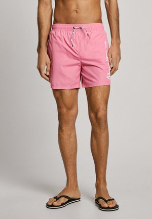 Bañador rosa hecho de material ligero, con cintura ajustable mediante cordón y detalle de logotipo en el lado. Combinado con chanclas negras.