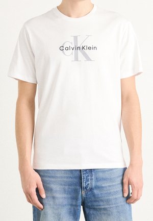 Mand iført en hvid Calvin Klein T-shirt med lyst gråt og sort logo kombineret med blå jeans, vist fra hals til hofter.