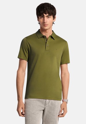 Giovane uomo con polo verde oliva a maniche corte e pantaloni grigio chiaro, in piedi davanti a uno sfondo semplice grigio chiaro.