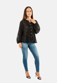 Blusa negra transparente con lunares blancos, cierre de botones en el frente, detalles con pliegues y mangas ligeramente abullonadas, combinada con jeans ajustados azules y tacones.