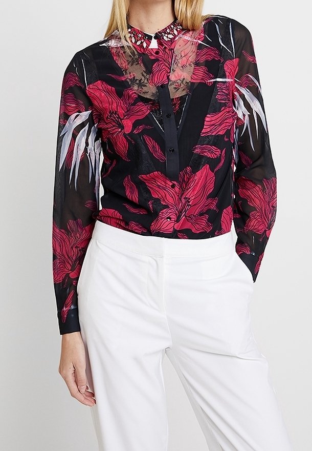 Schwarze transparente Bluse mit kräftigen roten Blumenmustern, Knöpfen an der Vorderseite und einem Spitzenkragen mit Verzierungen. Dazu weiße Hosen.