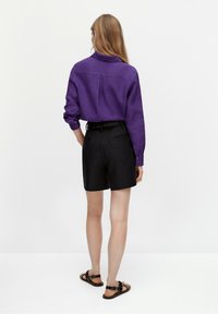 Femme aux cheveux longs portant une chemise à manches longues violette, un short noir ceinturé et des sandales noires, debout devant un fond clair uni.